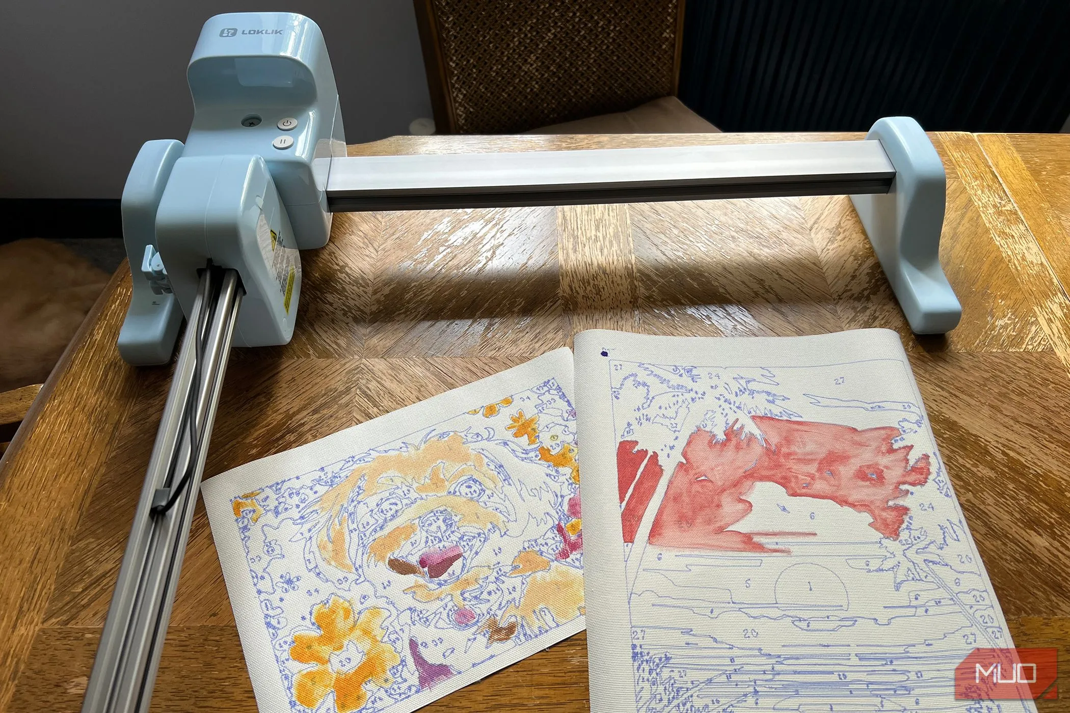 Hai bức tranh phong cảnh vẽ theo số đã hoàn thiện, đặt cạnh máy LOKLiK iPaint, minh họa kết quả nghệ thuật từ thiết bị AI