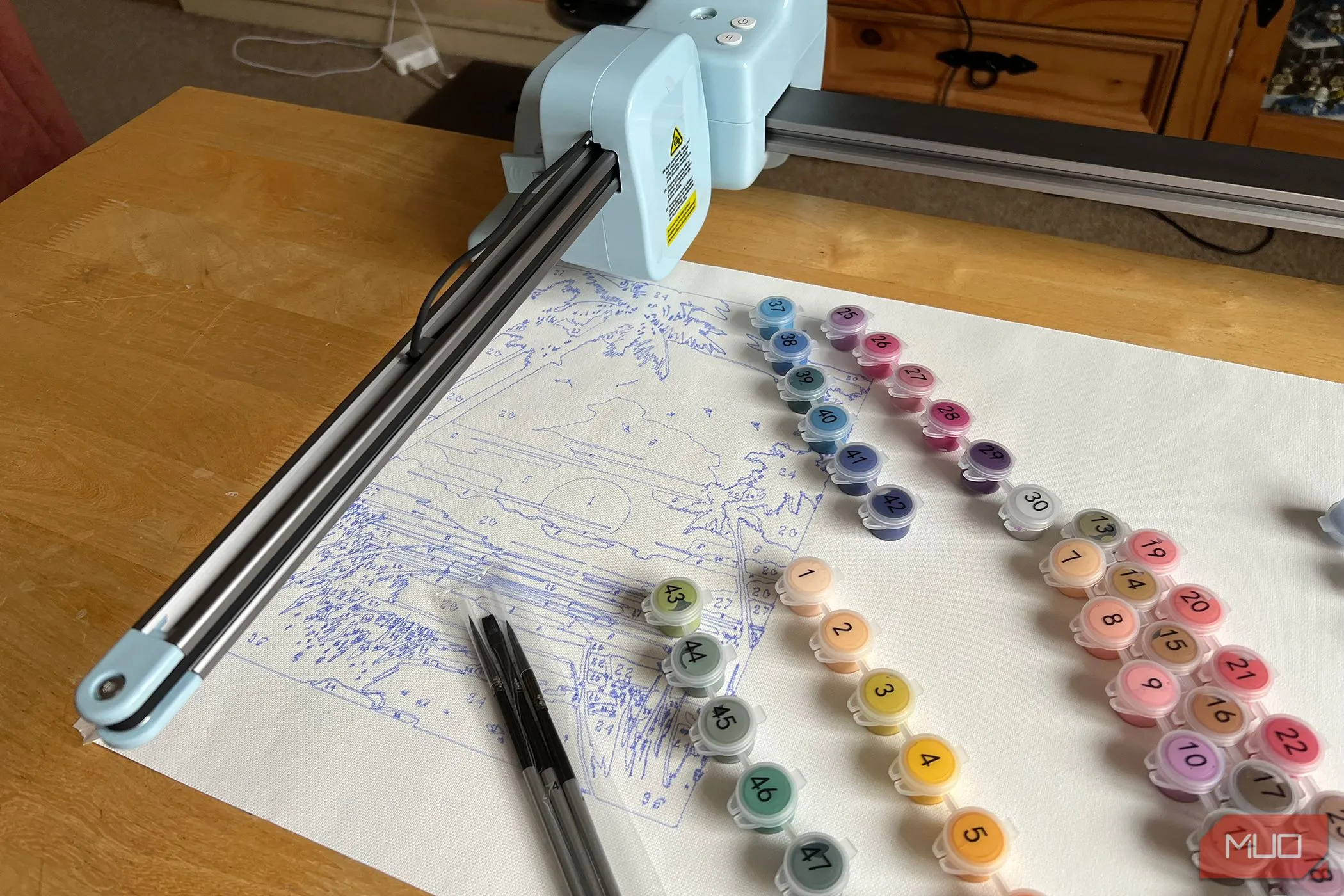 Máy LOKLiK iPaint đang vẽ một cảnh hoàng hôn trên canvas, với các hộp màu được đánh số đặt bên cạnh, sẵn sàng cho việc tô màu