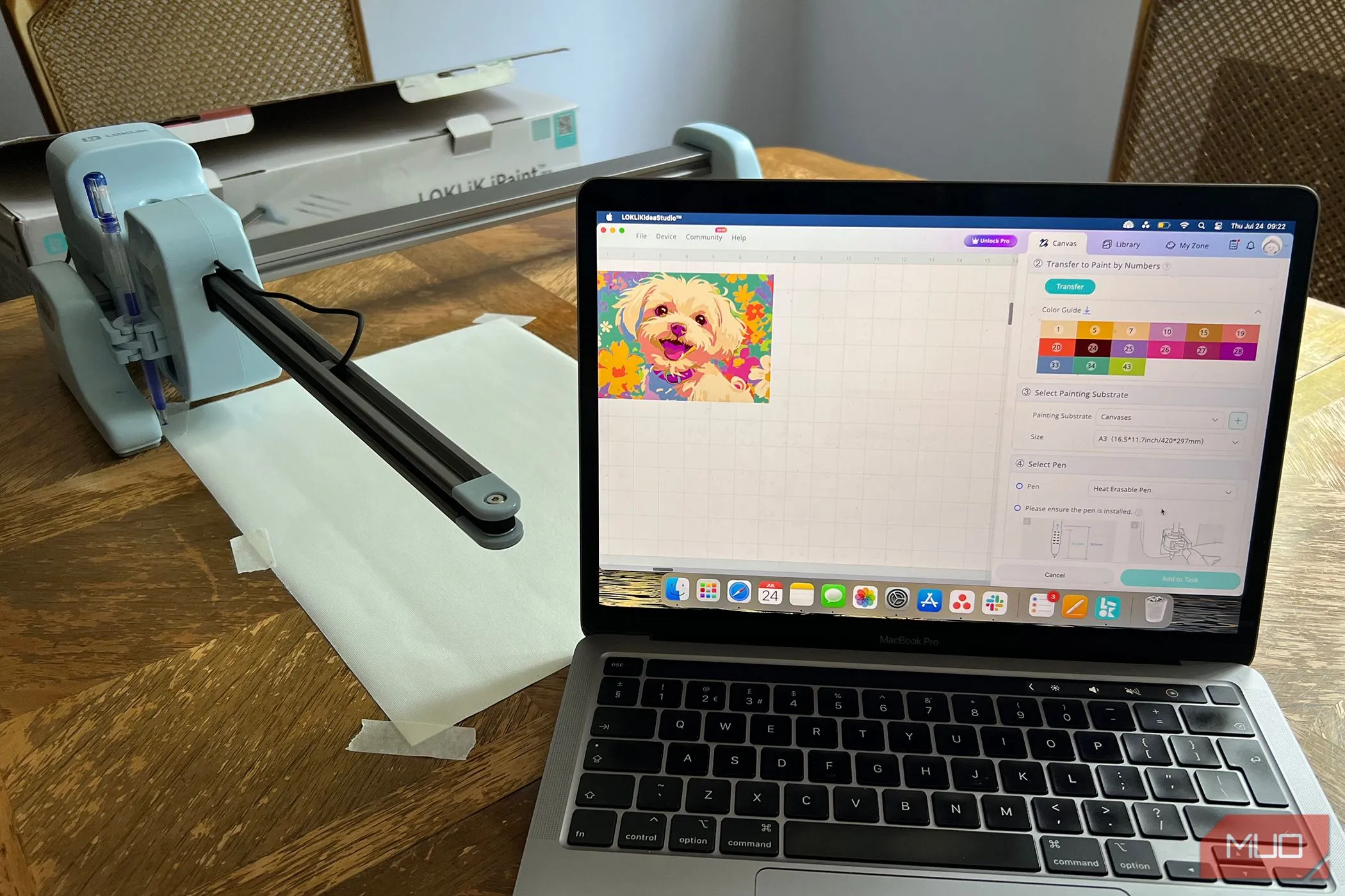 Máy LOKLiK iPaint đặt cạnh MacBook, hiển thị vải canvas trống và hình ảnh gốc trên màn hình chờ chuyển đổi thành tranh vẽ theo số